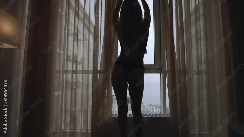 Silhouette of slim sexy woman opens a curtains at the morning in apartment with sea view