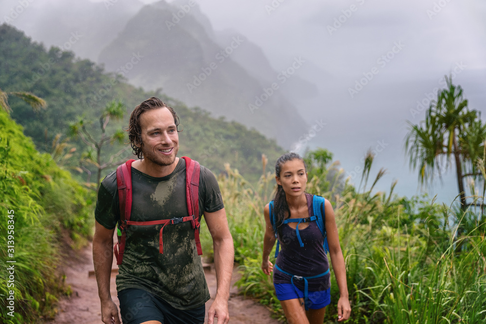 Mountain trek hikers couple walking in rain trekking on trail trek with backpacks healthy active lifestyle. Hiker man happy on hike in nature landscape with Asian girl, Na Pali Coast, Kauai, Hawaii.