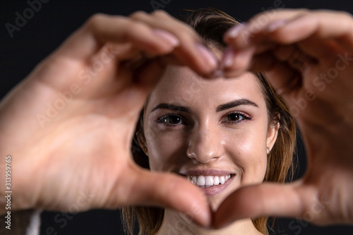 young lovely girl with a smile in an orange sweater shows a heart symbol with hands on a dark background