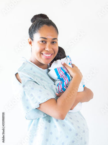 A smiling mixed race African American mother wearing a hospital gown holds her brand new infant baby on her shoulder hugging and cradling him as he sleeps in his blanket swaddle.