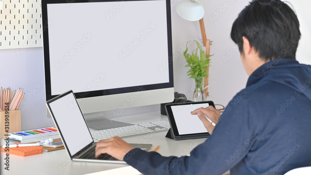 Back view of young creative man while using white blank screen laptop, tablet and computer at the working desk. Back view of young creative man while using white blank screen laptop, tablet and computer at the working desk.