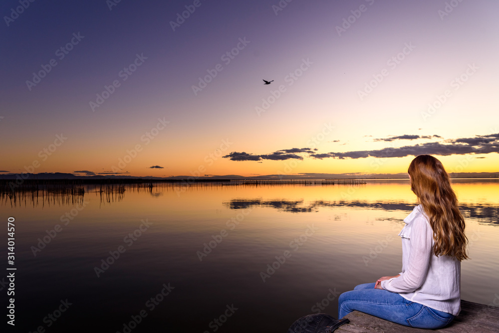 Long-haired young woman relaxing admiring the golden sunset on a beautiful pier by a natural lake. Long-haired young woman relaxing admiring the golden sunset on a beautiful pier by a natural lake.