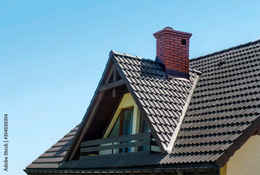 Roof with brown tiles, red chimney made by brick and attic with balcony against blue sky