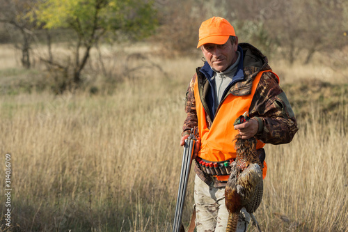 A man with a gun in his hands and an orange vest on a pheasant hunt in a wooded area in cloudy weather. A hunter with a pheasant in his hands.