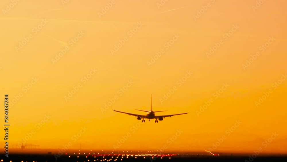 Back view silhouette of airplane landing at the airport at sunny orange and purple sunset. Runway end identification lights and strobe glowing. Back view silhouette of airplane landing at the airport at sunny orange and purple sunset. Runway end identification lights and strobe glowing.