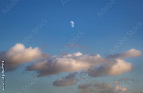 Half moon rising above fluffy clouds on a clear blue sky