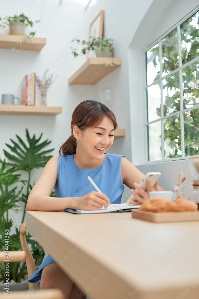 Smiling young Asian female entrepreneur sitting at a table and writing down notes Smiling young Asian female entrepreneur sitting at a table and writing down notes
