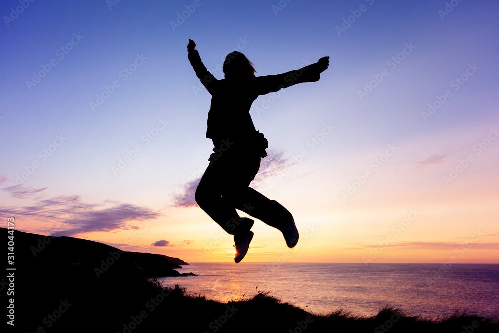 Silhouette of woman jumping for joy on coastal clifftops at sunset Silhouette of woman jumping for joy on coastal clifftops at sunset