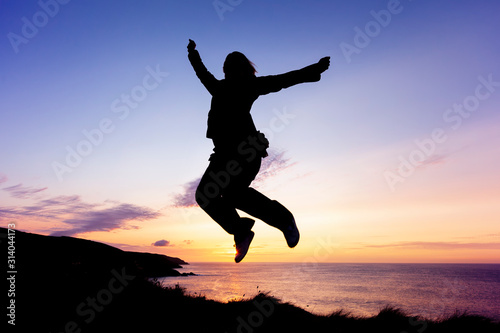 Silhouette of woman jumping for joy on coastal clifftops at sunset