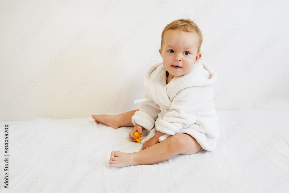pretty baby with a white bathrobe after taking a bath sitting on white cover on white background pretty baby with a white bathrobe after taking a bath sitting on white cover on white background