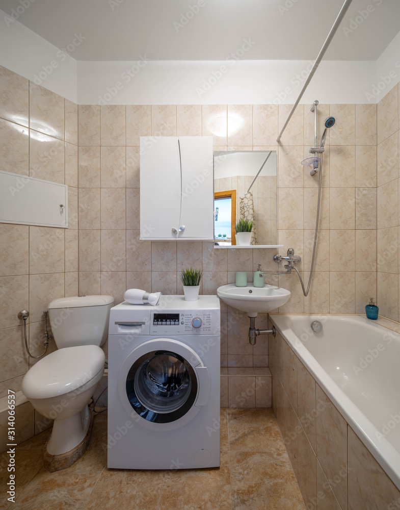 White toilet and sink with mirror. Bath and washing machine. Beige tile in bathroom. White toilet and sink with mirror. Bath and washing machine. Beige tile in bathroom.