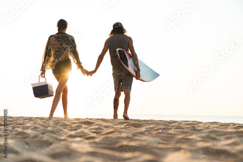 Photo from back of young couple holding surfboard and cooler bag