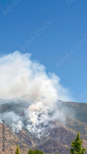 Vertical Mountain landscape with smoke from wild forest fire against clear blue sky