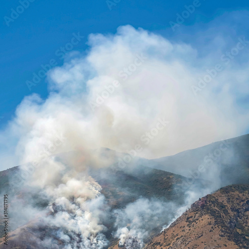 Square frame Aerial view of mountain with white smoke from wild forest fire on a sunny day © Jason