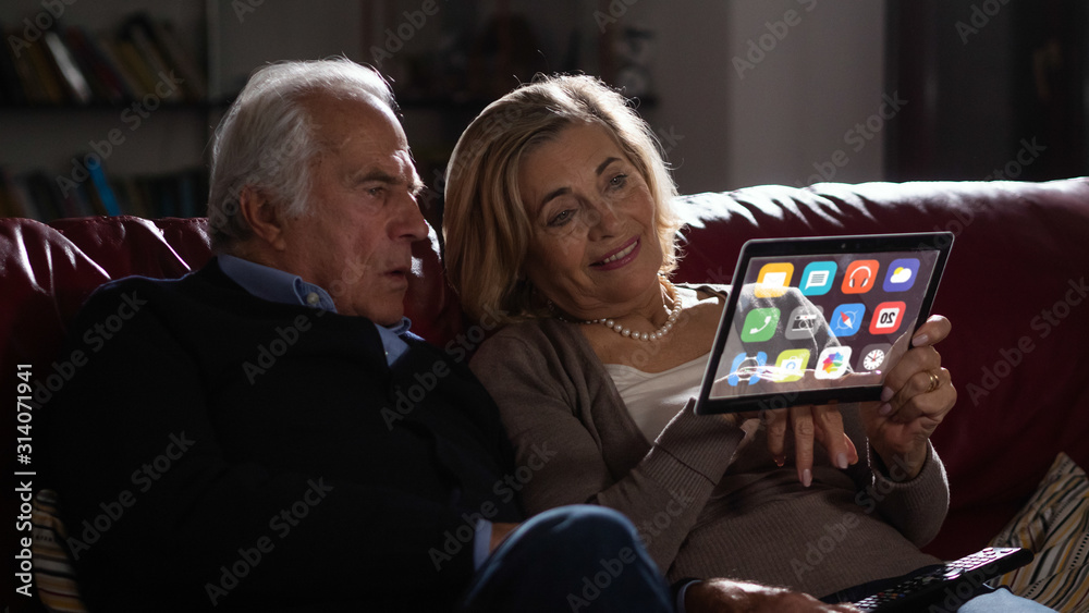 A modern senior couple is using a futuristic latest innovative technology glass tablet with augmented reality holograms on sofa in living room at home. A modern senior couple is using a futuristic latest innovative technology glass tablet with augmented reality holograms on sofa in living room at home.