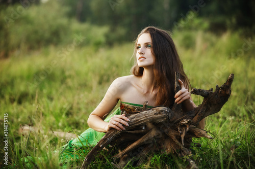 Girl in a beautiful green dress lies on the grass near the roots of a tree and dreams. Woman with bright makeup in nature, natural cosmetics