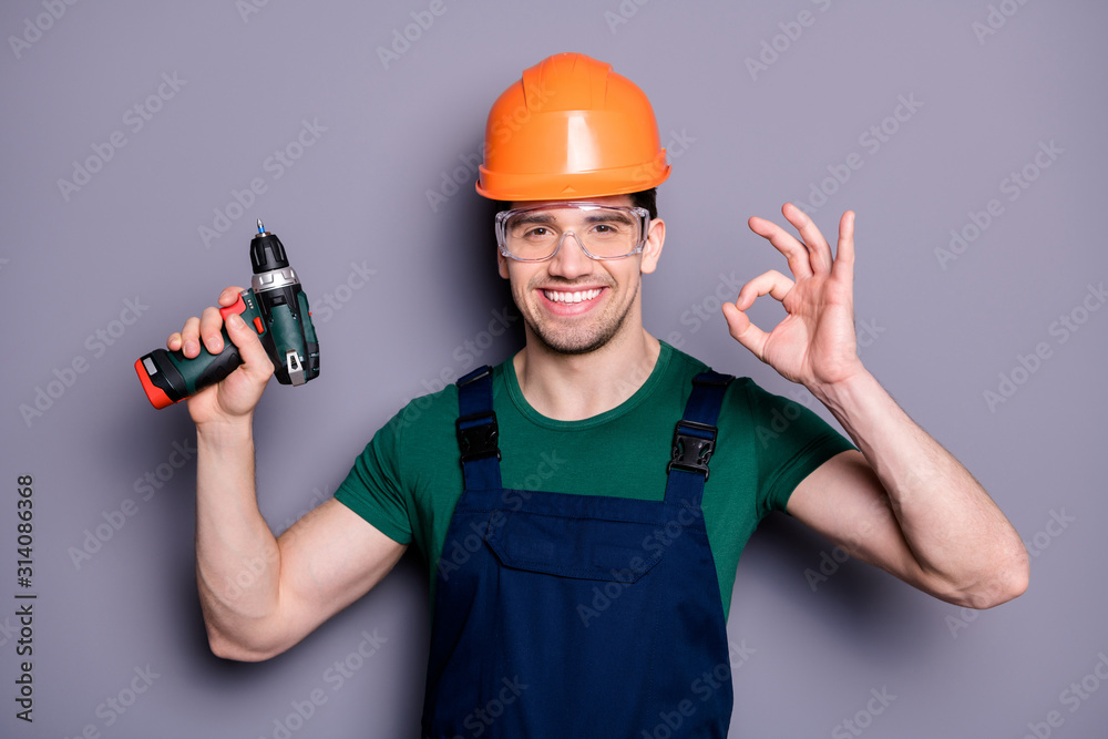 Photo of handsome masculine worker guy skilled engineer hold electric drill showing okey symbol wear t-shirt safety dungarees protective helmet glasses isolated grey background