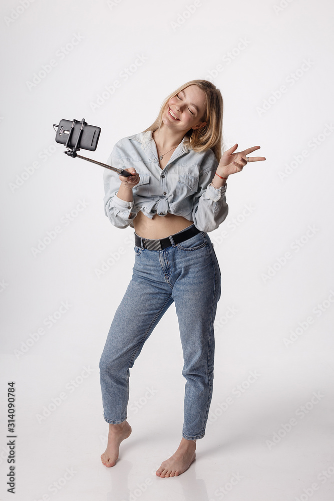 happy female making self portrait with smartphone attached to selfie stick  pole. young pretty blond girl wearing blue jeans smiling, using cellphone  isolated on white background. modern communication Stock Photo | Adobe