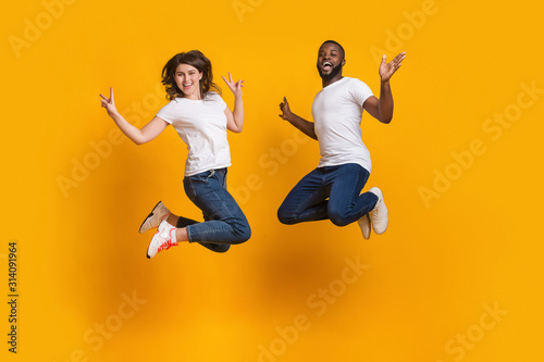 Excited interracial couple jumping, having fun together on yellow background