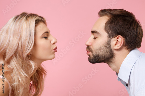 Side view of young couple two guy girl in party outfit celebrating posing isolated on pastel pink background in studio. People lifestyle Valentine's Day Women's Day birthday holiday concept. Kissing.