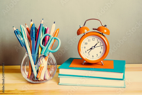 Orange alarm clock on books, handles, pencils, brushes in a glass on the table. School background 