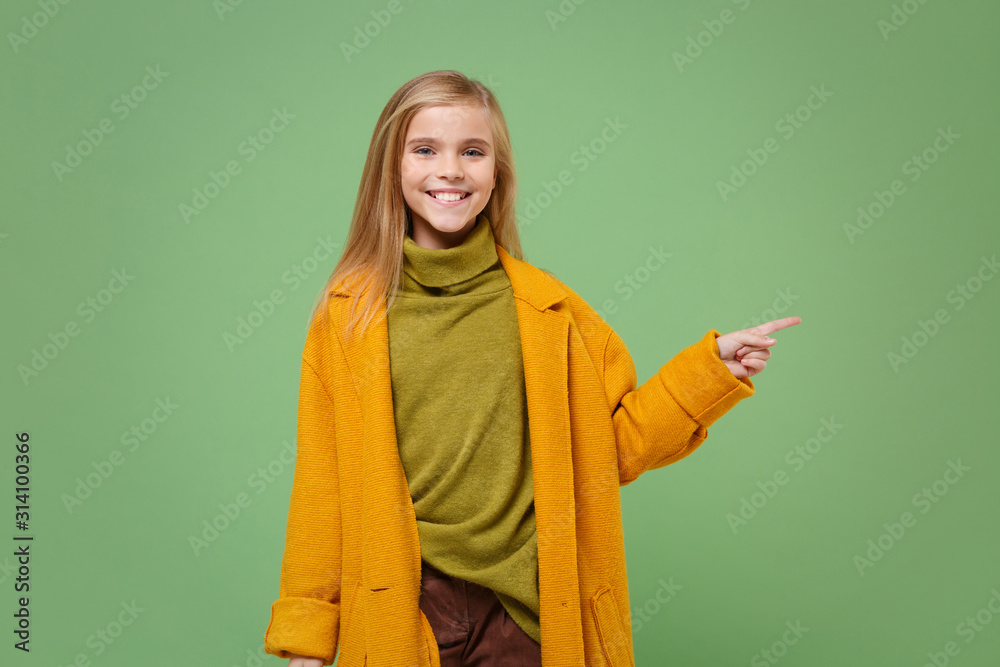 Smiling little blonde kid girl 12-13 years old in yellow coat posing isolated on pastel green background children portrait. Childhood lifestyle concept. Mock up copy space. Point index finger aside. Smiling little blonde kid girl 12-13 years old in yellow coat posing isolated on pastel green background children portrait. Childhood lifestyle concept. Mock up copy space. Point index finger aside.