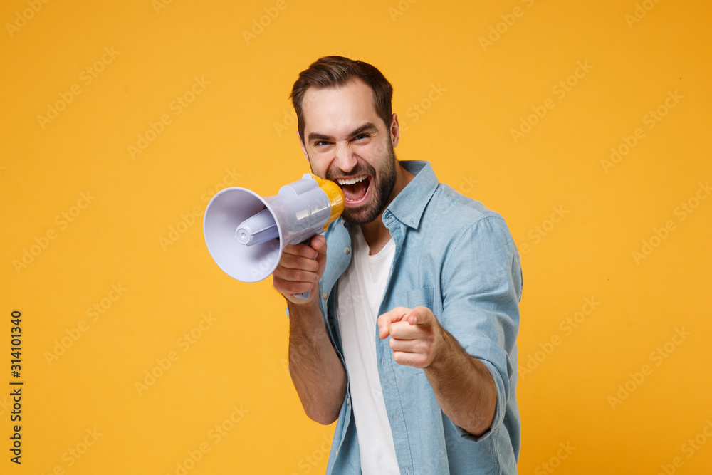 Funny young man in casual blue shirt posing isolated on yellow orange background studio portrait. People lifestyle concept. Mock up copy space. Screaming in megaphone pointing index finger on camera. - obrazy, fototapety, plakaty 