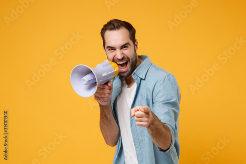 Funny young man in casual blue shirt posing isolated on yellow orange background studio portrait. People lifestyle concept. Mock up copy space. Screaming in megaphone pointing index finger on camera.