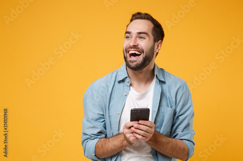 Laughing young man in casual blue shirt posing isolated on yellow orange wall background studio portrait. People emotions lifestyle concept. Mock up copy space. Using mobile phone, typing sms message.