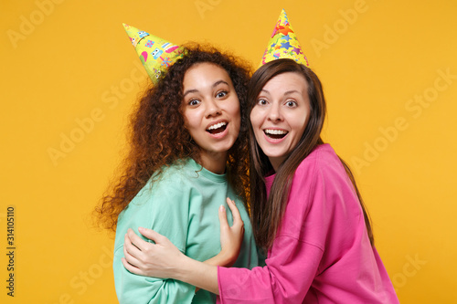Two excited women friends european and african american girls in pink green clothes, birthday hats posing isolated on yellow orange background. People lifestyle concept. Mock up copy space. Hugging.