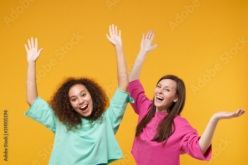 Two cheerful laughing women friends european and african american girls in pink green clothes posing isolated on yellow wall background. People lifestyle concept. Mock up copy space. Rising hands up.