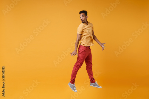 Shocked young african american guy in casual clothes posing isolated on yellow orange background studio portrait. People lifestyle concept. Mock up copy space. Jumping spreading hands looking aside.
