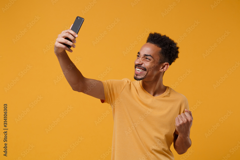 Happy young african american guy in casual t-shirt posing isolated on yellow orange background. People lifestyle concept. Mock up copy space. Doing selfie shot on mobile phone doing winner gesture. Happy young african american guy in casual t-shirt posing isolated on yellow orange background. People lifestyle concept. Mock up copy space. Doing selfie shot on mobile phone doing winner gesture.