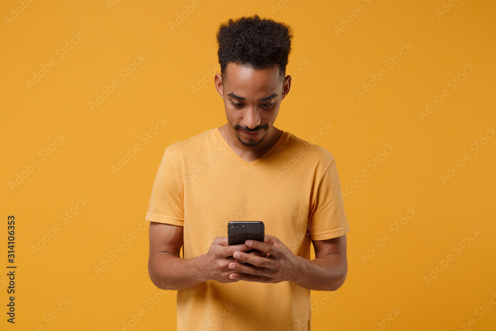 Young african american guy in casual t-shirt posing isolated on yellow orange wall background studio portrait. People lifestyle concept. Mock up copy space. Using mobile phone, typing sms message. Young african american guy in casual t-shirt posing isolated on yellow orange wall background studio portrait. People lifestyle concept. Mock up copy space. Using mobile phone, typing sms message.
