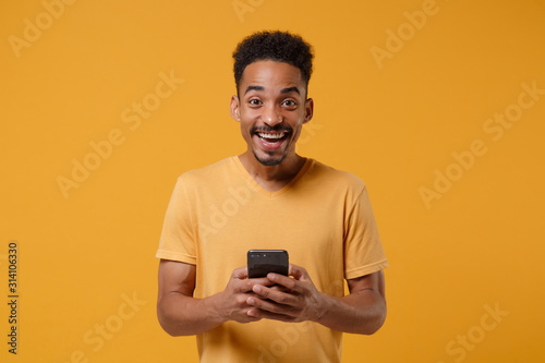 Laughing young african american guy in casual t-shirt posing isolated on yellow orange background studio portrait. People lifestyle concept. Mock up copy space. Using mobile phone, typing sms message.