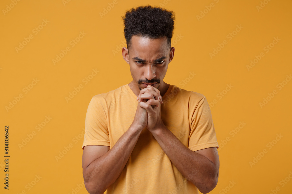 Young african american guy in casual t-shirt posing isolated on yellow orange wall background studio portrait. People emotions lifestyle concept. Mock up copy space. Holding hands folded in prayer. Young african american guy in casual t-shirt posing isolated on yellow orange wall background studio portrait. People emotions lifestyle concept. Mock up copy space. Holding hands folded in prayer.