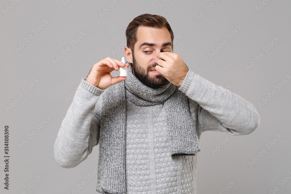 Young man in gray sweater, scarf isolated on grey background studio portrait. Healthy lifestyle ill sick disease treatment cold season concept. Mock up copy space. Hold nasal drops put hand on nose. Young man in gray sweater, scarf isolated on grey background studio portrait. Healthy lifestyle ill sick disease treatment cold season concept. Mock up copy space. Hold nasal drops put hand on nose.
