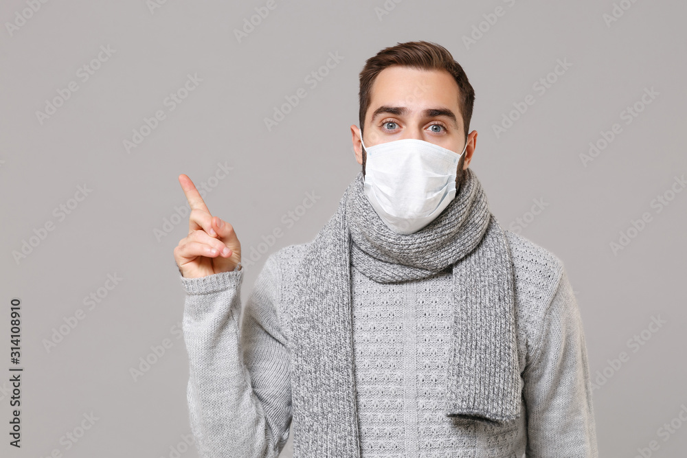 Young man in gray sweater, scarf isolated on grey background. Healthy lifestyle, ill disease treatment, cold season concept. Mock up copy space. Wearing sterile face mask, point index finger aside up. Young man in gray sweater, scarf isolated on grey background. Healthy lifestyle, ill disease treatment, cold season concept. Mock up copy space. Wearing sterile face mask, point index finger aside up.