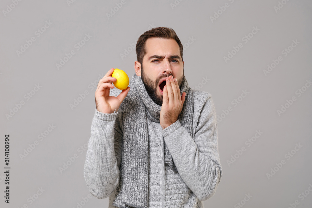 Young man in gray sweater, scarf posing isolated on grey background in studio. Healthy lifestyle, ill sick disease treatment, cold season concept. Mock up copy space. Hold lemon, coughing, sneezing. Young man in gray sweater, scarf posing isolated on grey background in studio. Healthy lifestyle, ill sick disease treatment, cold season concept. Mock up copy space. Hold lemon, coughing, sneezing.