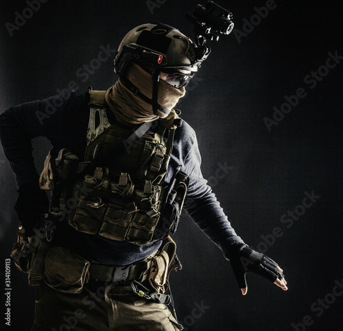 Special operations fighter in helmet with night-vision  thermal imaging device  load carrier carefully moving with caution in darkness  holding hand on pistol  ready for fight during dangerous mission