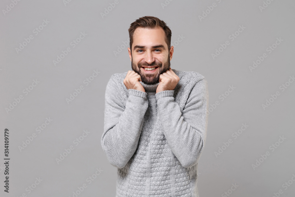 Smiling frozen young bearded man in gray sweater posing isolated on grey background studio portrait. Healthy fashion lifestyle people emotions cold season concept. Mock up copy space. Clenching fists. Smiling frozen young bearded man in gray sweater posing isolated on grey background studio portrait. Healthy fashion lifestyle people emotions cold season concept. Mock up copy space. Clenching fists.