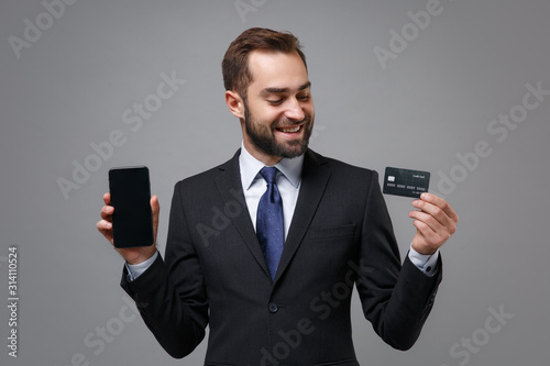 Funny business man in suit shirt tie posing isolated on grey background. Achievement career wealth business concept. Mock up copy space. Holding mobile phone with blank empty screen, credit bank card.