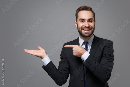 Smiling young bearded business man in classic black suit shirt tie posing isolated on grey background. Achievement career wealth business concept. Mock up copy space. Pointing index finger hand aside.