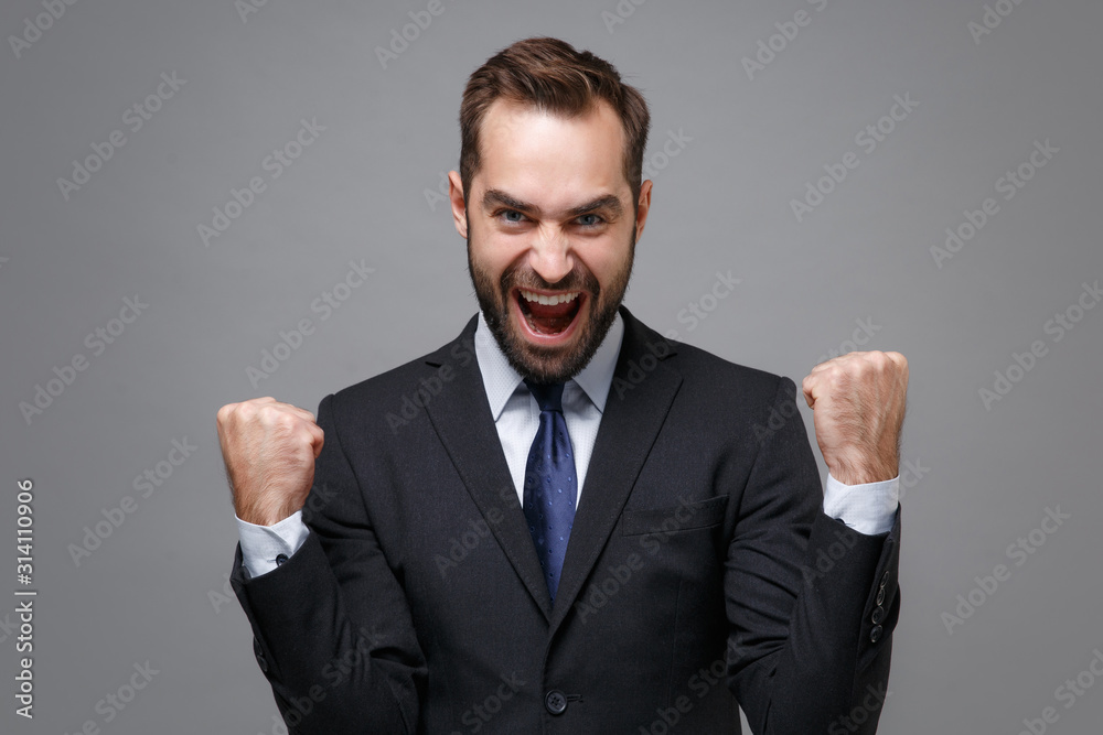 Crazy joyful young business man in classic black suit shirt tie posing isolated on grey wall background in studio. Achievement career wealth business concept. Mock up copy space. Doing winner gesture. Crazy joyful young business man in classic black suit shirt tie posing isolated on grey wall background in studio. Achievement career wealth business concept. Mock up copy space. Doing winner gesture.