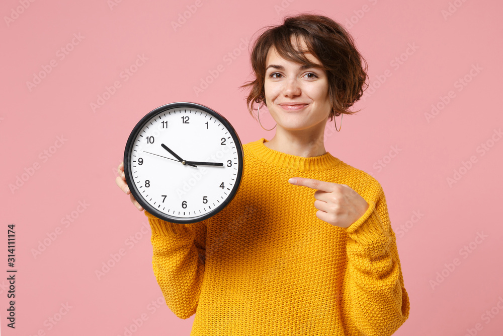 Smiling young brunette woman girl in yellow sweater posing isolated on pastel pink background in studio. People sincere emotions lifestyle concept. Mock up copy space. Pointing index finger on clock. - obrazy, fototapety, plakaty 