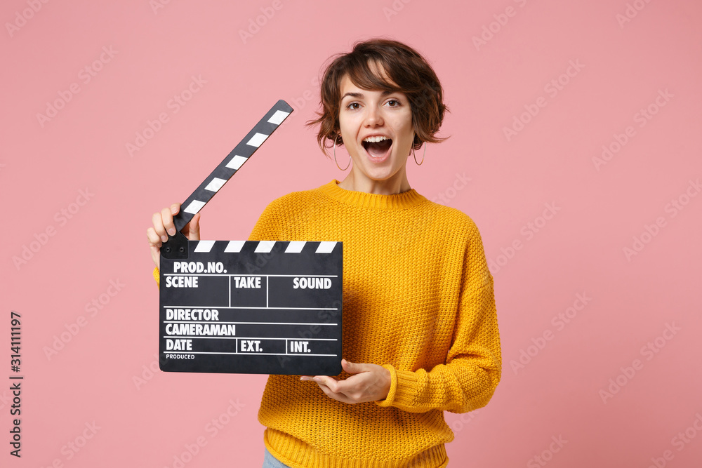 Excited young brunette woman girl in yellow sweater posing isolated on pastel pink background in studio. People lifestyle concept. Mock up copy space. Holding classic black film making clapperboard.