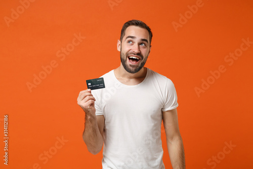 Cheerful young man in casual white t-shirt posing isolated on orange background studio portrait. People sincere emotions lifestyle concept. Mock up copy space. Holding credit bank card, looking up.