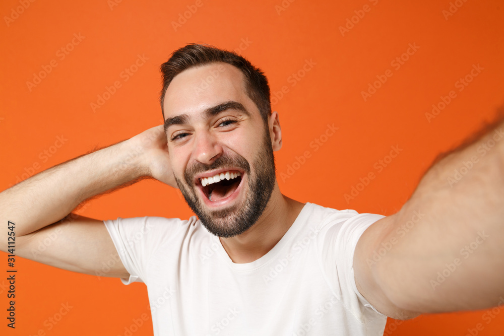 Close up of cheerful funny young man in casual white t-shirt posing isolated on orange wall background studio portrait. People lifestyle concept. Mock up copy space. Doing selfie shot on mobile phone. Close up of cheerful funny young man in casual white t-shirt posing isolated on orange wall background studio portrait. People lifestyle concept. Mock up copy space. Doing selfie shot on mobile phone.