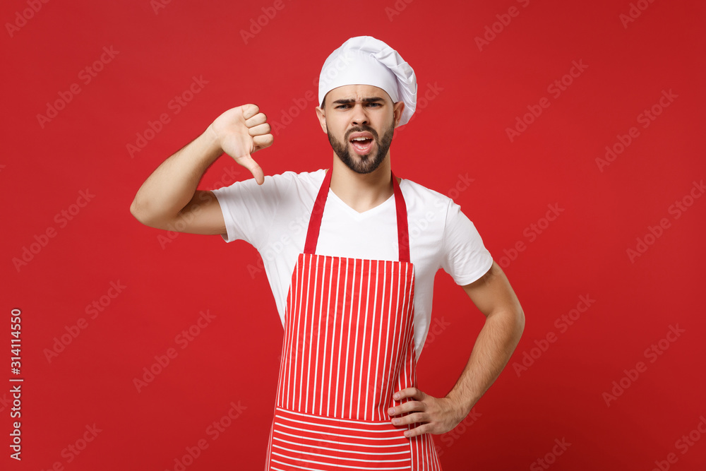 Displeased young bearded male chef cook or baker man in striped apron white t-shirt toque chefs hat posing isolated on red background. Cooking food concept. Mock up copy space. Showing thumb down. Displeased young bearded male chef cook or baker man in striped apron white t-shirt toque chefs hat posing isolated on red background. Cooking food concept. Mock up copy space. Showing thumb down.