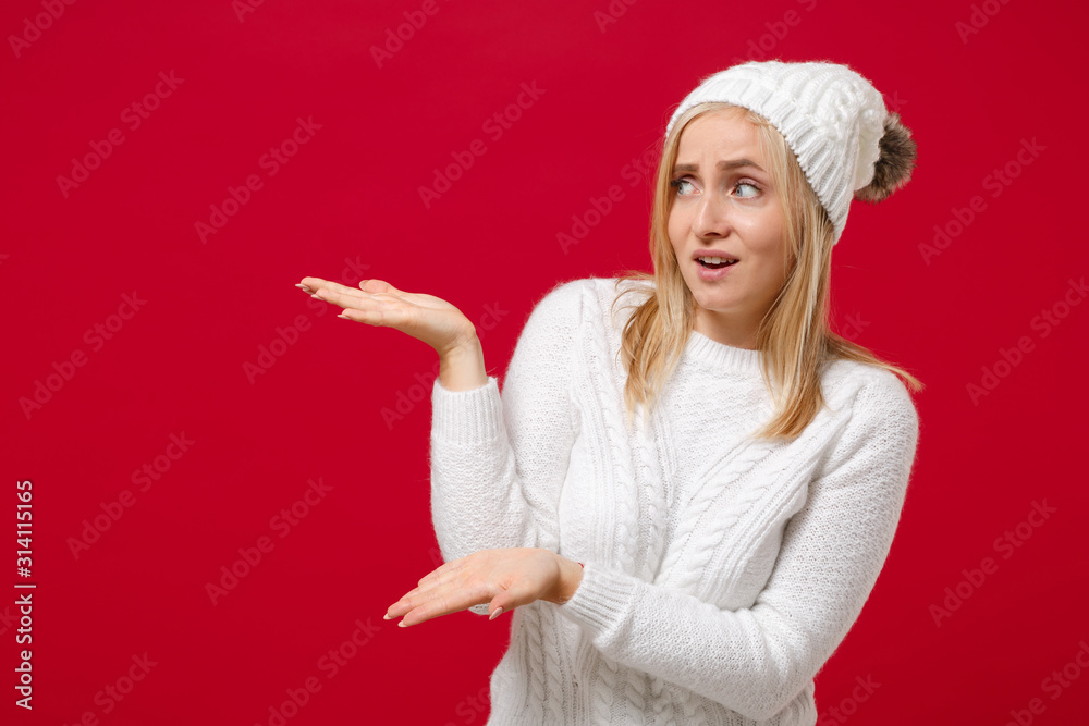 Bewildered young woman in white sweater hat isolated on red wall background studio portrait. Healthy fashion lifestyle people emotions cold season concept. Mock up copy space. Pointing hands aside. Bewildered young woman in white sweater hat isolated on red wall background studio portrait. Healthy fashion lifestyle people emotions cold season concept. Mock up copy space. Pointing hands aside.